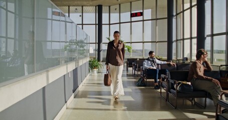An elegant young woman walks through the lobby at the airport. People are waiting for a flight sitting in chairs