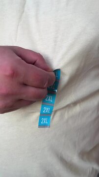 Vertical 4K video. Fat adult man in light beige T-shirt removes blue sticker with text 2XL. Concept of buying new clothes of large size, Black Friday sale, shopping in mall, fitting, weight loss. Shop