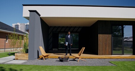 The businessman is near the house. He smiles at the camera. A sunny summer day