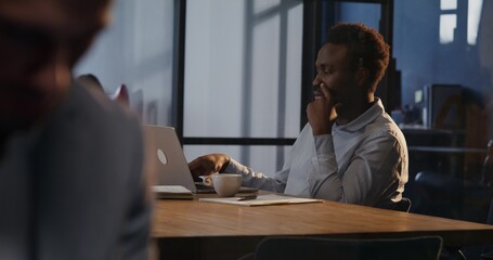 A man works in an office. He uses a laptop while sitting at his desk with a serious look