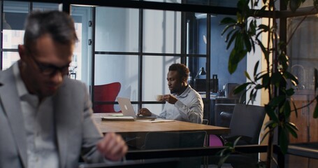 A man works in the office next to his colleague. He uses a laptop and drinks coffee sitting at the table.