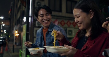 Happy young man and woman laughing and talking standing on a city street at night. They're eating takeaways.