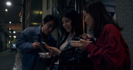 Young people use a mobile phone standing on the street at night city. They eat street food and smile happily.