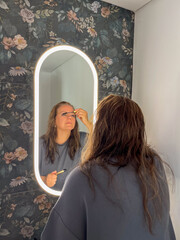 Midlife woman applying mascara in front of a floral mirror, showcasing beauty routine and...