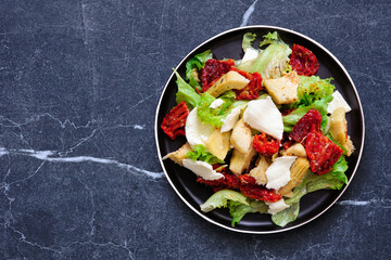 Italian salad with fresh mozzarella cheese, sun dried tomatoes, artichokes and lettuce. Top view on a dark stone background.
