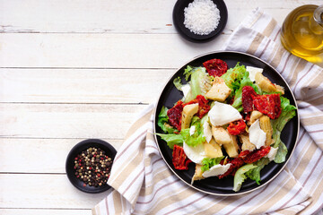 Italian salad with fresh mozzarella cheese, sun dried tomatoes, artichokes and lettuce. Top down view table scene on a white wood background.