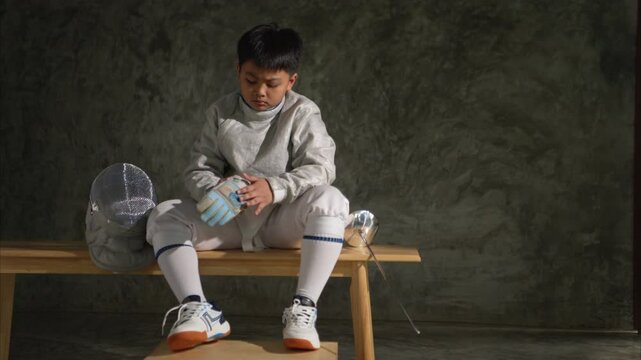 Young boy in fencing gear sitting on a bench, examining his &eacute;p&eacute;e sword with focus. Helmet placed beside him