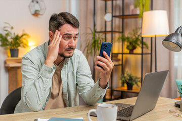Upset displeased Caucasian man freelancer using smartphone sitting at table desk in living room....