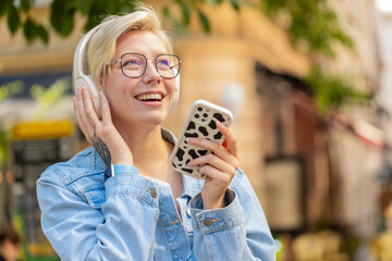 Happy relaxed overjoyed young blonde girl in wireless headphones choosing, listening favorite...