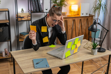 Tired frustrated exhausted young businessman sits at home office table with laptop sticky notes...