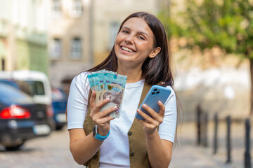 Happy woman counting UAH Ukrainian hryvnias money cash using smartphone calculator app satisfied of...
