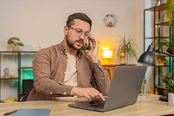 Young businessman wearing headset, freelance worker, call center or support service operator helpline, talking with client or colleague communication support. Guy using laptop at home office desk.