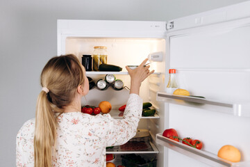 Woman long blonde hair, wearing floral blouse, is reaching into refrigerator filled with fresh...