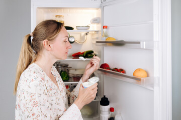 Woman enjoying yogurt while standing in front of an open refrigerator filled with fresh fruits and...