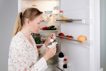 Woman enjoying yogurt from a container while standing in front of an open refrigerator filled with...