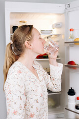 Woman enjoying refreshing glass of water in the morning, standing in front of open refrigerator...