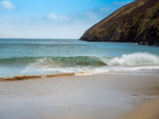 Ocean wave hits sandy beach, rough coastline with cliffs and blue cloudy sky in the background. Stunning Keem bay and beach in county Mayo, Ireland. Travel and tourism. Irish nature landscape.