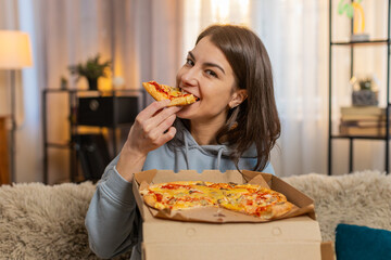 Happy Caucasian young woman receives pizza delivery sitting on sofa with the box quickly unpacks...