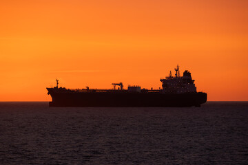 The dark silhouette of a large crude oil tanker ship sailing across the open ocean during a spectacular and vibrant sunset.
