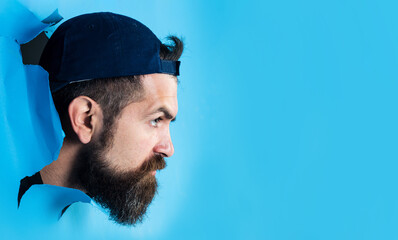 Serious bearded man breaking through paper wall. Handsome male in cap looking through hole in...
