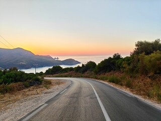 Fototapeta premium a winding coastal road in Annaba, Algeria, at sunset. The sky transitions from warm oranges to soft blues, illuminating the Sea. Vegetation frames the road, leading the eye towards the serene waters.
