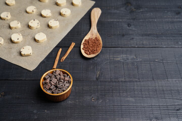 Front view of cookie dough pieces on baking paper with wooden spoon of cacao, chocolate chips bowl...