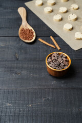 Vertical front view of cut cookie dough on baking paper with wooden spoon filled with cacao, bowl of chocolate chips and cinnamon sticks