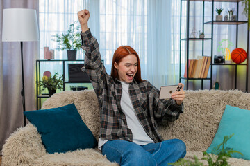 Young woman at home playing mobile game with excitement tapping on screen and celebrating happy...