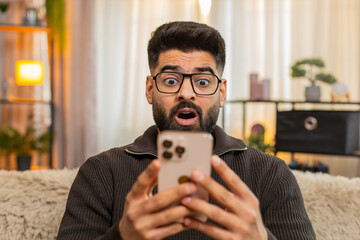 Indian man at home opens eyes wide and lifts eyebrows while looking at smartphone with shocked...