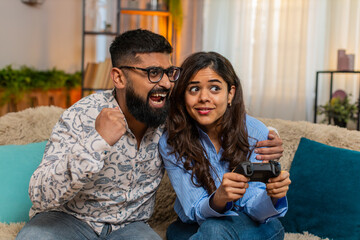 Indian couple man and woman sitting on home sofa with game controller and popcorn, laughing and...