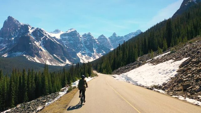 Cyclist riding toward Moraine Lake on a scenic mountain road surrounded by snowy peaks and dense pine forest. Concept of freedom, eco-adventure, and breathtaking Canadian landscapes.