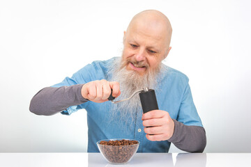 Elderly man with a white beard joyfully using a hand-crank coffee grinder, dressed in a blue shirt with gray cuffs, isolated on a white backdrop