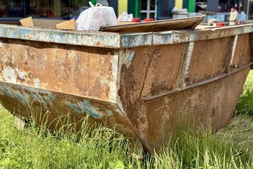 An Old and Rusty Dump Container That is Surrounded by Lush, Overgrown Grass and Weeds. A weathered metal dumpster overflowing with various debris, positioned against a backdrop of lush tall grass.