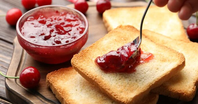 Woman making toast with tasty cherry jam at wooden table, closeup
