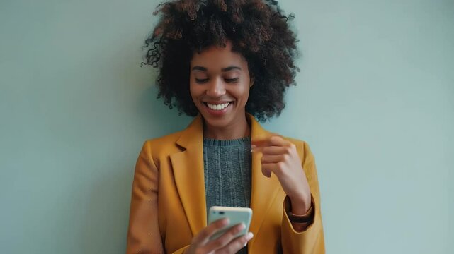 Smiling person with curly hair looking at a smartphone against a teal wall wearing a yellow blazer and gray sweater. She appears happy while using her phone indoors.