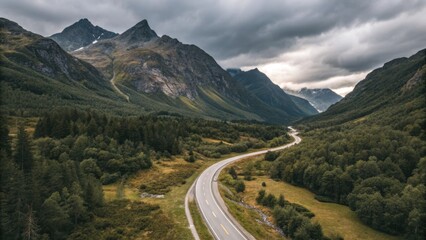 Fototapeta premium Winding highway cuts through a majestic valley with mountains and lush forests, underneath a moody, dramatic sky