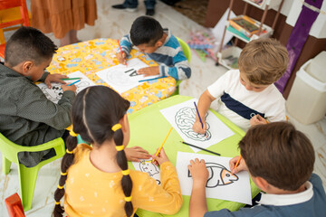 Preschool children sit around a table coloring under their teacher’s guidance. One kindergarten...