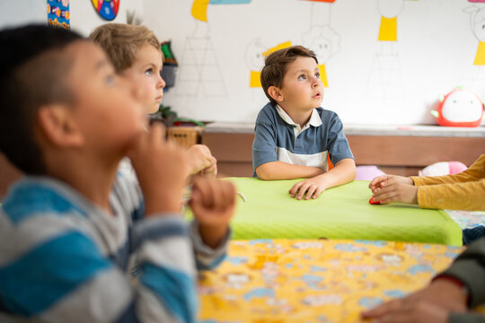 Preschool children sitting around a table listening attentively, with one boy looking up in deep thought. Kindergarten learning environment focused on attention, curiosity, and early education