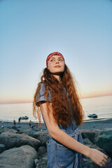 young woman with long curly hair wearing a red bandana and denim outfit enjoying sunset at rocky...