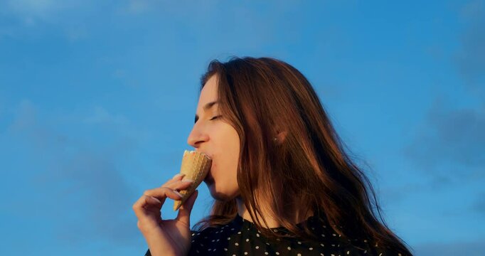 Woman savoring ice cream cone against blue sky, woman enjoys cool vanilla flavor as cone melts in sunlight, scene continues with breeze through hair and sweet cream dripping down cone tip.