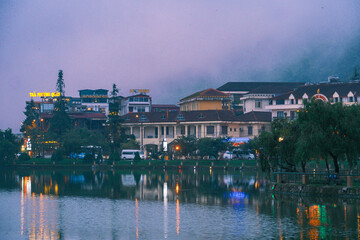 Naklejka premium Sapa city lake at night with misty reflections and illuminated buildings creating a serene travel destination vibe, 사파 시내 호수 야경과 안개 낀 반영, 불빛으로 연출된 여행지 분위기
