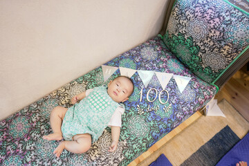 A 100-day-old Japanese baby lies comfortably on a vibrant, colorful sofa, with festive decorations...