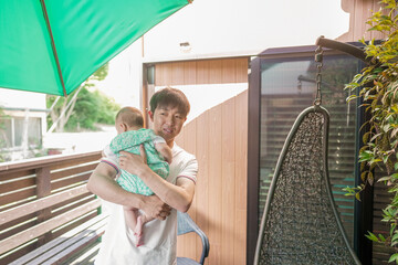 A Japanese father in his thirties holds his 100-day-old baby on a sunlit terrace with potted...