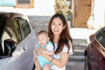 In bright sunshine, a Japanese mother in her twenties stands in her home's driveway garage, holding...