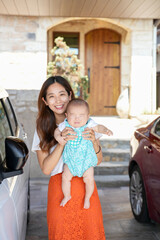 In bright sunshine, a Japanese mother in her twenties stands in her home's driveway garage, holding...