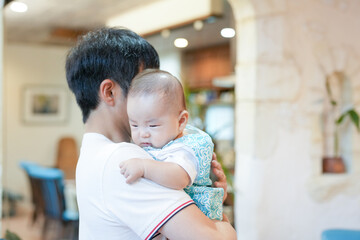 In a bright living room, a Japanese father in his thirties sits on a red sofa cradling his...