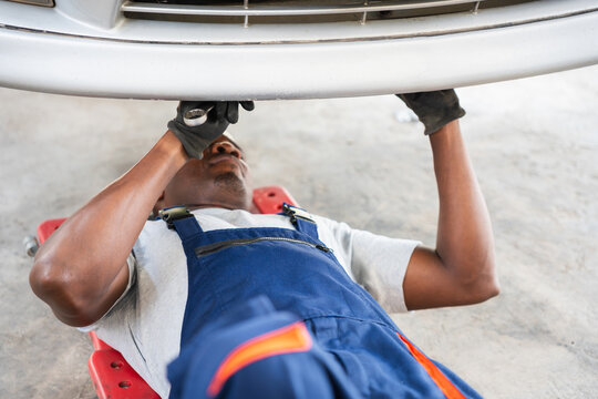 African American Mechanic Working Under Car on Creeper, Male Technician Inspecting Undercarriage of Vehicle, Professional Mechanic Diagnosing Car Problems in Repair Shop