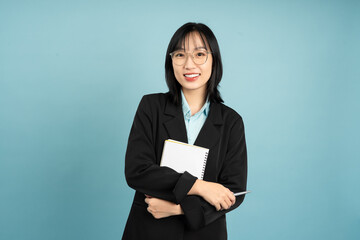 Young Asian professional poses confidently against a solid blue background while holding a notebook...