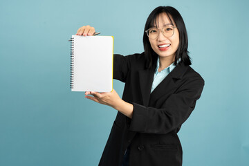 Smiling woman in a black blazer holds a blank notebook while posing against a blue background,...