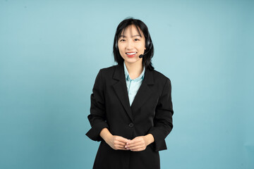 Professional Asian woman wearing a black suit with a headset standing against a blue background...